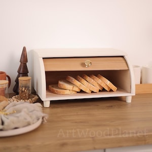 Puede incluir: Una panera de madera blanca y natural con puerta enrollable y pomo dorado. La panera está llena de pan en rebanadas. La panera está sobre una superficie de madera. El texto "ArtWoodPlanet" es visible.