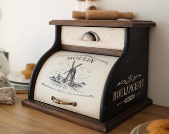 May include: A wooden bread bin with a dark brown frame and a cream-coloured roll-top door. The door features a black graphic of a windmill with the text MOULIN DEPUIS 1852. The side is labelled BOULANGERIE. It includes a top drawer with a metal handle.