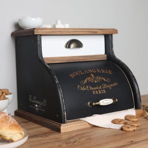 Op de afbeelding: Een zwart-witte houten broodtrommel met een roldeur voorzien van de gouden tekst BOULANGERIE, Cafe Chaud et Beignets, PARIS. De unit bevat een bovenste lade met een bronzen handgreep en een onderste deur met een witte keramische handgreep.