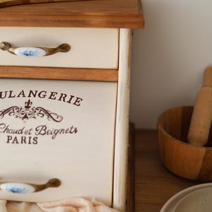 Puede incluir: Una panera de madera de color crema con una puerta frontal abatible que presenta el texto marr&oacute;n "BOULANGERIE" y "Chaud et Beignets PARIS". La unidad incluye un caj&oacute;n superior con un tirador de cer&aacute;mica blanca con motivos florales azules.