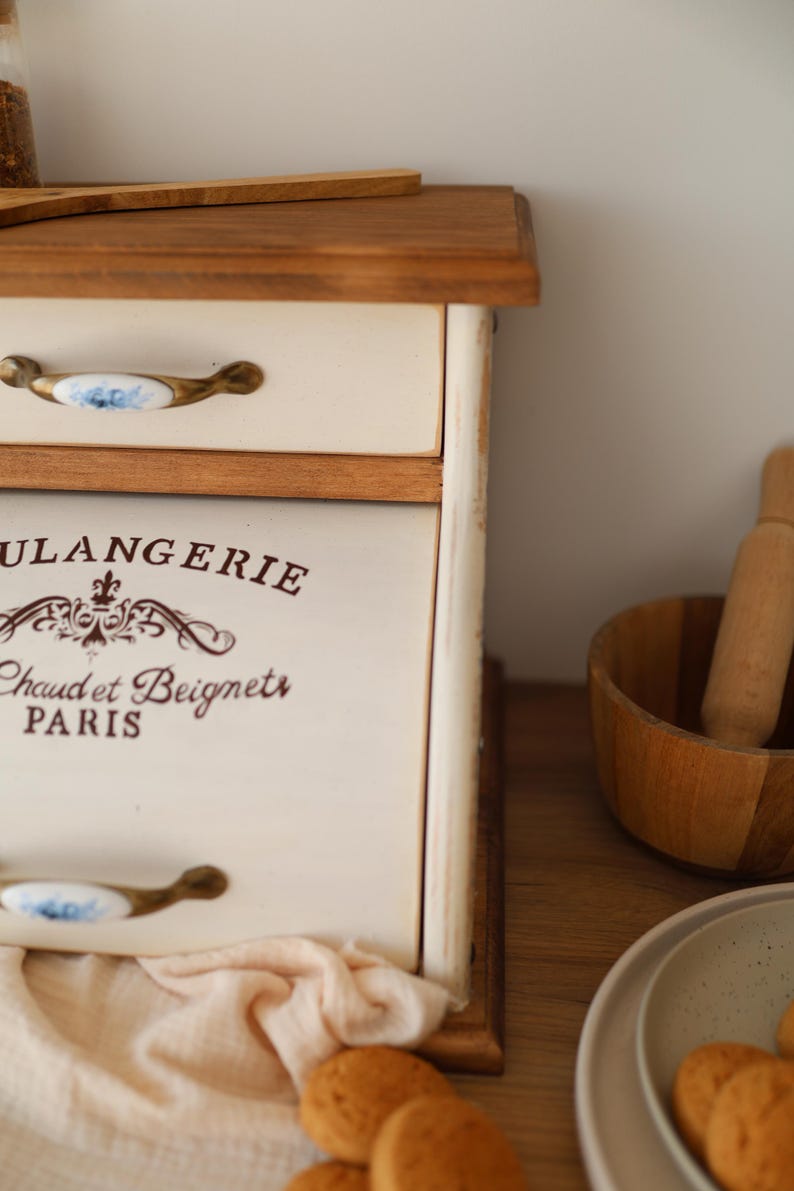 Puede incluir: Una panera de madera de color crema con una puerta frontal abatible que presenta el texto marr&oacute;n "BOULANGERIE" y "Chaud et Beignets PARIS". La unidad incluye un caj&oacute;n superior con un tirador de cer&aacute;mica blanca con motivos florales azules.