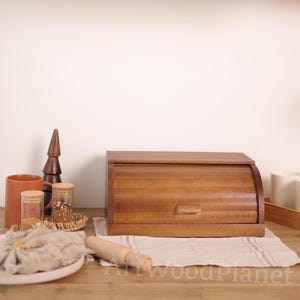 Könnte beinhalten: Eine Holz-Brotdose mit Rolldeckel, aus warm getöntem Holz gefertigt. Die Box steht auf einer Holzoberfläche, zusammen mit einem Nudelholz, einem Teller mit einer Serviette und anderem Küchenzubehör. Die Gesamtästhetik ist rustikal und einladend.