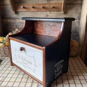 May include: A wooden bread box featuring a black top and side panels, a white front drawer with a brown knob, and the text "La Boulangerie" and "Patisserie" printed on the exterior. The design includes a Paris emblem on the interior back panel.