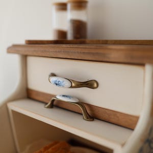 Puede incluir: Una panera de madera con un panel frontal de color crema y dos cajones con tiradores de lat&oacute;n antiguo con inserciones de cer&aacute;mica blanca decoradas con motivos florales azules. Varias rebanadas de pan son visibles en el compartimento inferior.
