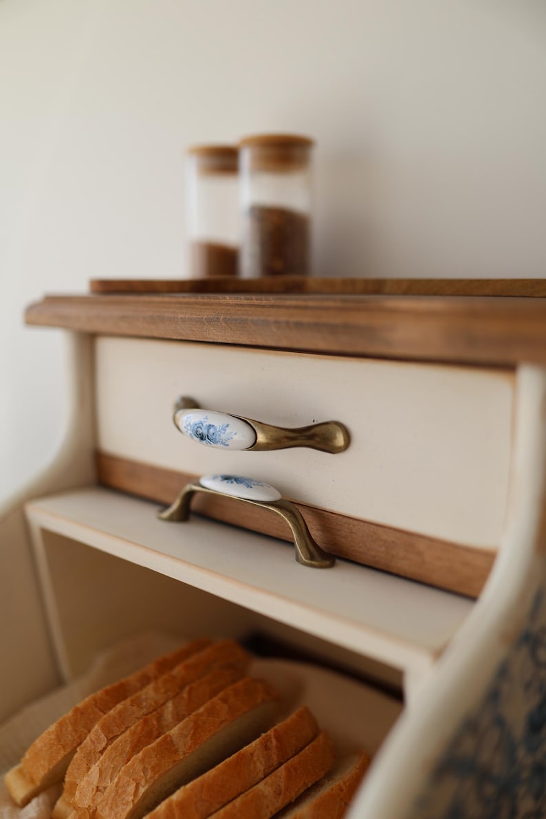 Puede incluir: Una panera de madera con un panel frontal de color crema y dos cajones con tiradores de lat&oacute;n antiguo con inserciones de cer&aacute;mica blanca decoradas con motivos florales azules. Varias rebanadas de pan son visibles en el compartimento inferior.