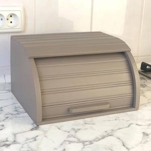 May include: A taupe-colored wooden bread box with a roll-top door and a handle. The box has a curved top and is sitting on a marble countertop. The bread box is designed to keep bread fresh.