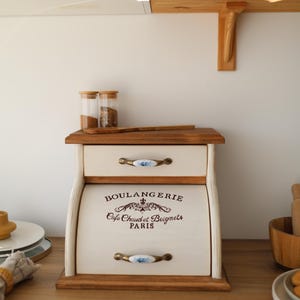 Puede incluir: Una panera de madera con acabado en color crema, que cuenta con un caj&oacute;n superior y una puerta enrollable. La puerta muestra el texto BOULANGERIE, Cafe Chaud et Beignets, PARIS en marr&oacute;n oscuro. Tiene dos tiradores de metal con detalles florales azules.
