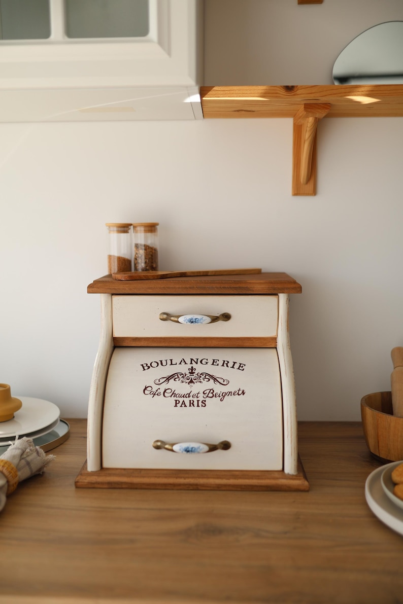 Puede incluir: Una panera de madera con acabado en color crema, que cuenta con un caj&oacute;n superior y una puerta enrollable. La puerta muestra el texto BOULANGERIE, Cafe Chaud et Beignets, PARIS en marr&oacute;n oscuro. Tiene dos tiradores de metal con detalles florales azules.