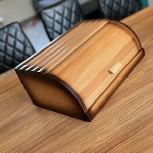 Puede incluir: Una panera de madera con la parte superior curvada y un peque&ntilde;o tirador de madera. La panera est&aacute; hecha de madera de color claro con bordes marrones oscuros y se encuentra sobre una mesa de madera. La panera est&aacute; dise&ntilde;ada para mantener el pan fresco.