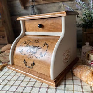 May include: A wooden bread box with a natural wood finish and cream-colored sides. It features a roll-top door with the black text Boulangerie de Paris, a small top drawer with a black knob, and a decorative metal handle on the front door.