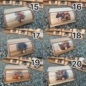 Op de afbeelding: Houten brooddozen met diverse bloemmotieven. Elke doos heeft een gebogen deksel en een natuurlijke houten afwerking. De ontwerpen omvatten zonnebloemen, lavendel, bessen en tarwe. De dozen worden tentoongesteld op een bed van kleine grijze stenen.