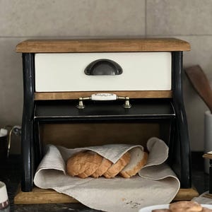 Puede incluir: Una panera de madera con un caj&oacute;n blanco y detalles negros. La parte superior e inferior son de madera natural. La panera est&aacute; abierta, revelando rebanadas de pan sobre un pa&ntilde;o de lino. Un tirador decorativo est&aacute; en el caj&oacute;n.
