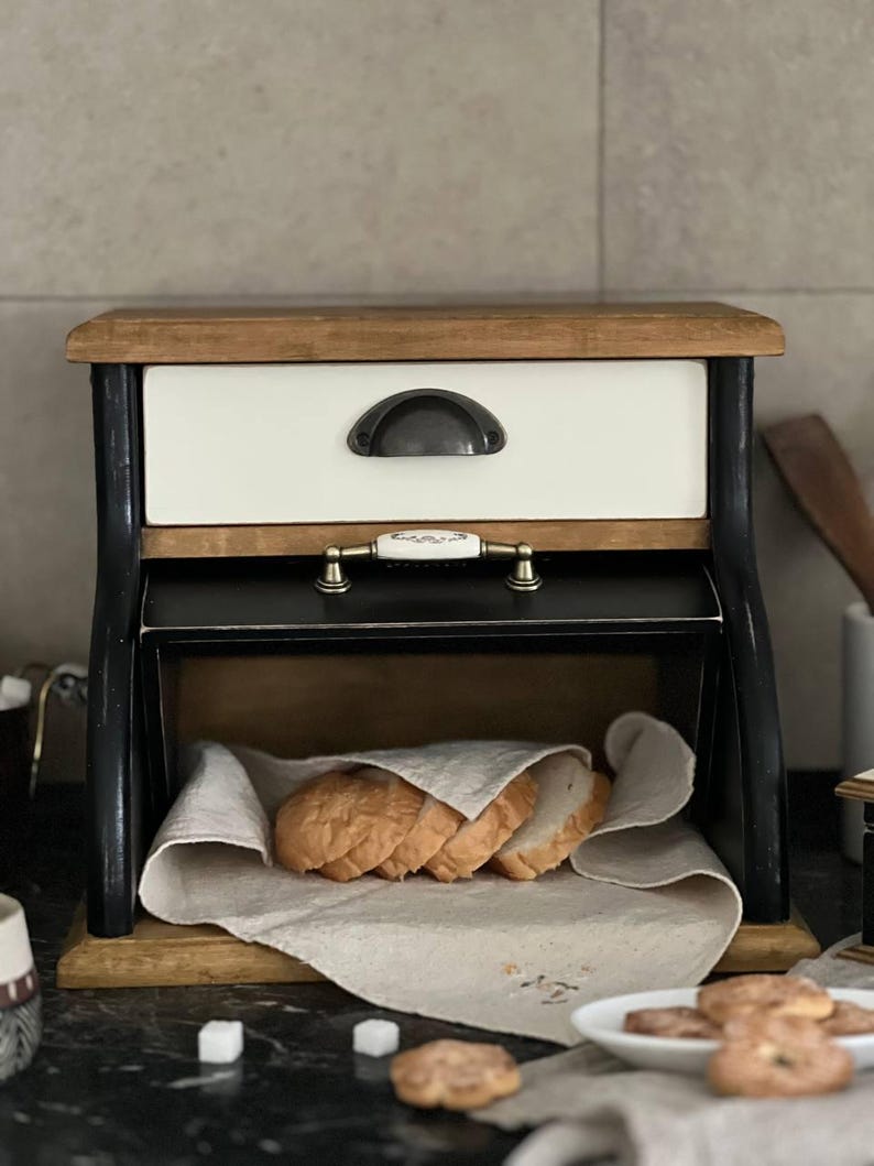 Puede incluir: Una panera de madera con un caj&oacute;n blanco y detalles negros. La parte superior e inferior son de madera natural. La panera est&aacute; abierta, revelando rebanadas de pan sobre un pa&ntilde;o de lino. Un tirador decorativo est&aacute; en el caj&oacute;n.