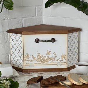Puede incluir: Una panera de esquina de madera con tapa marrón oscuro y cuerpo blanco. La parte delantera presenta un diseño decorativo de pájaros y flores. Los lados tienen un patrón de celosía. Un asa de metal está centrada en la parte delantera. Pan en rodajas y un cuchillo en primer plano.