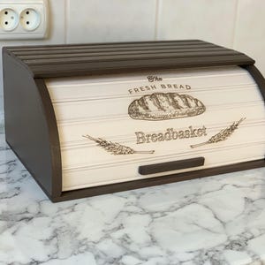 May include: A wooden breadbox with a roll-top lid. The box is brown with a cream-colored front panel. The front panel has the words "The Fresh Bread" and "Breadbasket" with a bread illustration and wheat stalks.