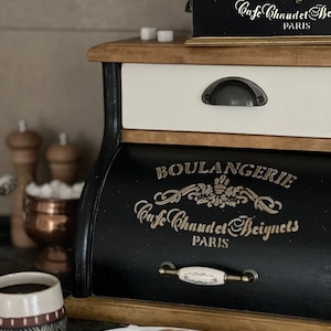 Puede incluir: Una panera de madera en blanco y negro con la inscripci&oacute;n dorada "BOULANGERIE Cafe Chaudet Beignets PARIS". Un plato blanco peque&ntilde;o con galletas y terrones de az&uacute;car est&aacute; delante. Tambi&eacute;n se ven una taza de caf&eacute; y una peque&ntilde;a caja negra.