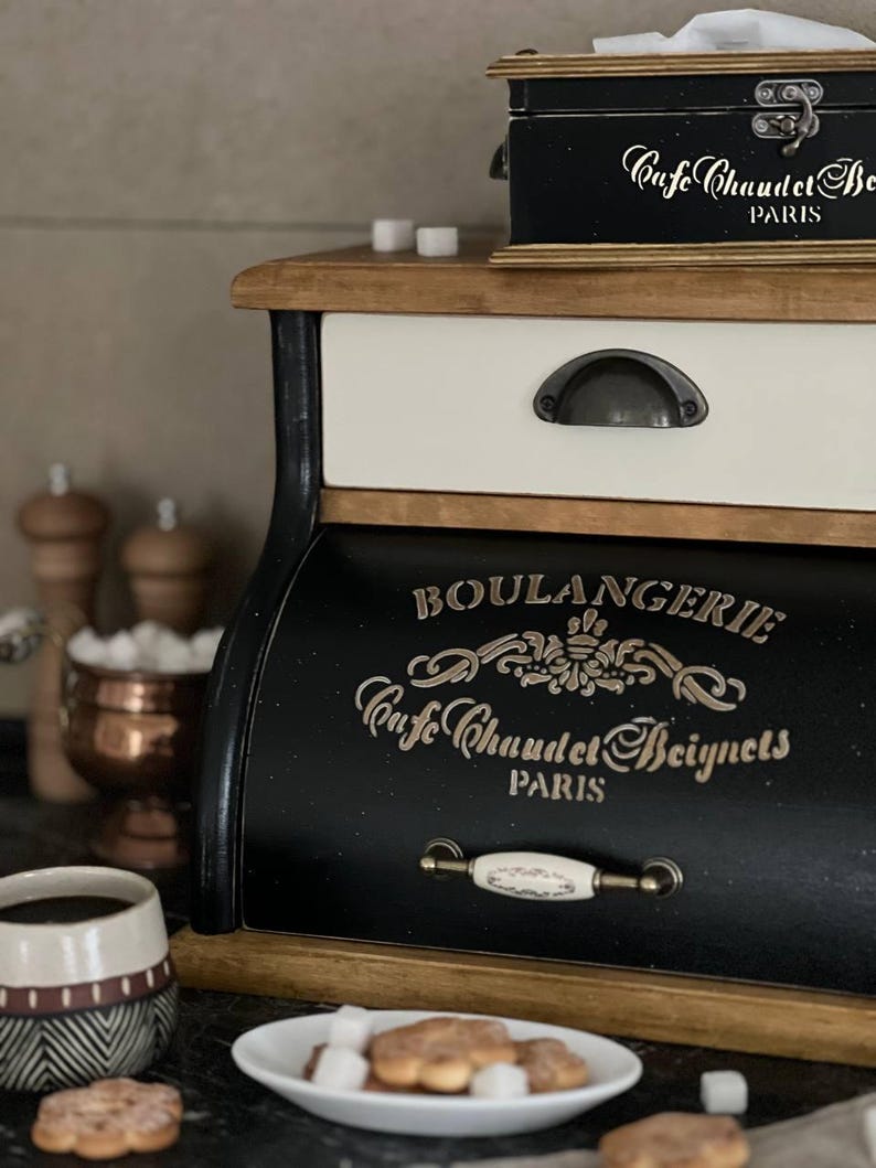Puede incluir: Una panera de madera en blanco y negro con la inscripci&oacute;n dorada "BOULANGERIE Cafe Chaudet Beignets PARIS". Un plato blanco peque&ntilde;o con galletas y terrones de az&uacute;car est&aacute; delante. Tambi&eacute;n se ven una taza de caf&eacute; y una peque&ntilde;a caja negra.