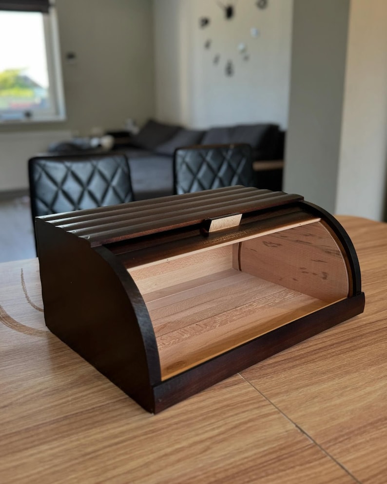 Puede incluir: Una panera de madera marr&oacute;n oscuro con un dise&ntilde;o curvo. La caja tiene una tapa de listones y un interior de color claro. La panera est&aacute; sobre una mesa de madera. El tema general es el almacenamiento de cocina.
