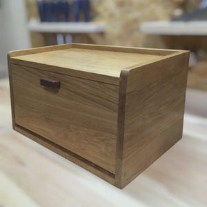 Pode incluir: Uma caixa de pão retangular de madeira clara. Possui uma parte superior plana, uma gaveta retrátil e uma pequena alça marrom escura. A caixa de pão é feita de madeira maciça e apresenta um grão natural.
