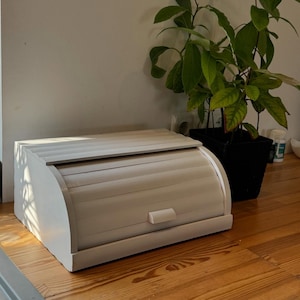 Puede incluir: Una panera de madera blanca con tapa enrollable. La panera tiene una base rectangular y una parte superior curva. Una peque&ntilde;a manija rectangular est&aacute; en la parte delantera. Una planta en maceta con hojas verdes est&aacute; al lado de la panera.