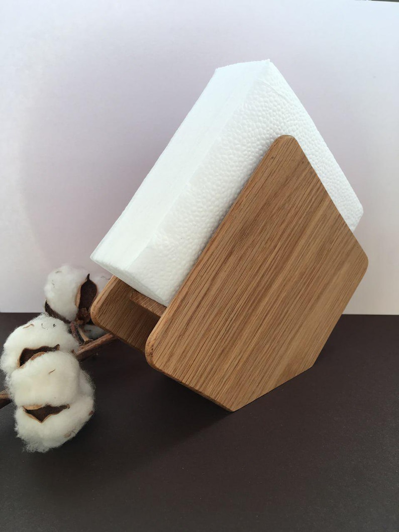 Puede incluir: Un servilletero de madera con un dise&ntilde;o geom&eacute;trico, que sostiene una pila de servilletas blancas. El soporte est&aacute; hecho de madera clara y tiene un patr&oacute;n de vetas natural. Una ramita de algod&oacute;n a&ntilde;ade un toque decorativo.