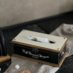 Puede incluir: Un dispensador de pa&ntilde;uelos rectangular con base negra y tapa crema. La caja tiene un marco de madera y presenta el texto "Cafe Chaudet Beignets PARIS" en escritura blanca. La caja est&aacute; sobre un pa&ntilde;o de lino con galletas y cubos de az&uacute;car.