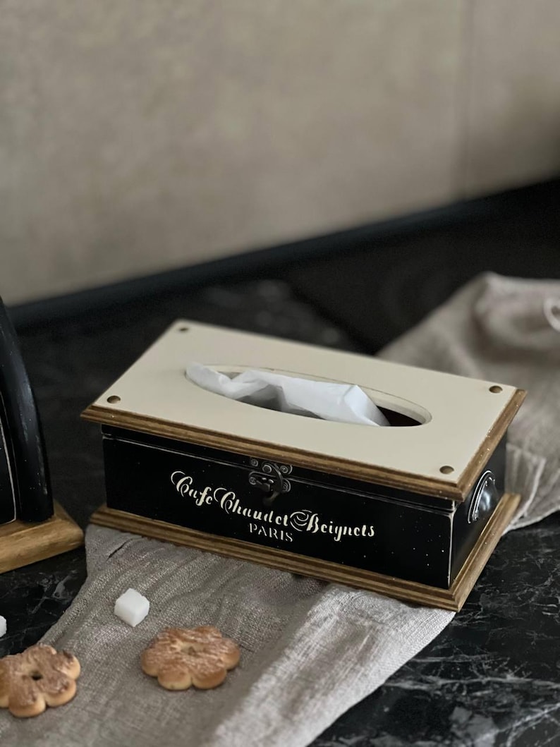 Puede incluir: Un dispensador de pa&ntilde;uelos rectangular con base negra y tapa crema. La caja tiene un marco de madera y presenta el texto "Cafe Chaudet Beignets PARIS" en escritura blanca. La caja est&aacute; sobre un pa&ntilde;o de lino con galletas y cubos de az&uacute;car.