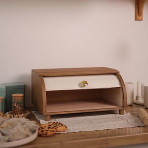 Puede incluir: Panera de madera con tapa enrollable. La panera tiene un acabado de madera clara con un frente crema y un pomo dorado. La panera está sobre una superficie de madera.