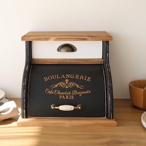 May include: A wooden bread box with a natural wood top and base, a white drawer with a bronze cup pull, and a black roll-top door featuring gold text that reads BOULANGERIE, Cafe Chaud et Beignets, PARIS, and a horizontal white handle.
