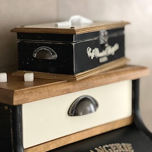 Puede incluir: Una caja de pa&ntilde;uelos negra y de madera con ribete dorado y un asa de metal se encuentra encima de un peque&ntilde;o caj&oacute;n blanquecino con un asa de metal. El caj&oacute;n forma parte de un mueble m&aacute;s grande con la palabra "BOULANGERIE" en letras doradas.