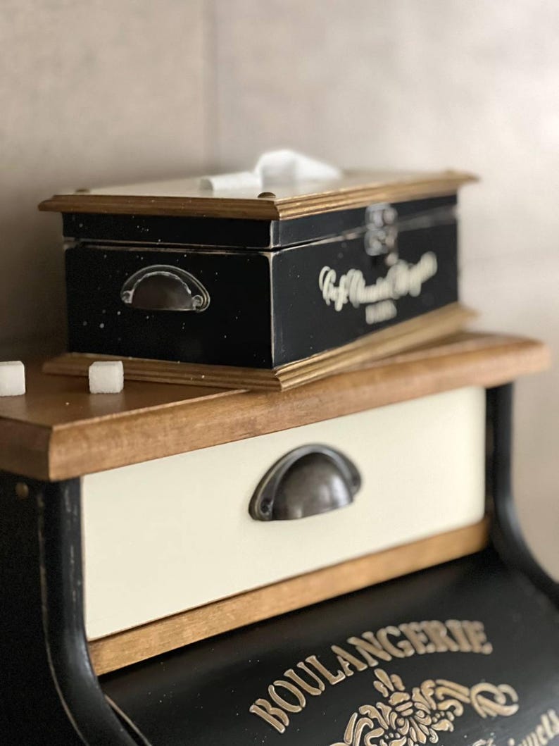 Puede incluir: Una caja de pa&ntilde;uelos negra y de madera con ribete dorado y un asa de metal se encuentra encima de un peque&ntilde;o caj&oacute;n blanquecino con un asa de metal. El caj&oacute;n forma parte de un mueble m&aacute;s grande con la palabra "BOULANGERIE" en letras doradas.