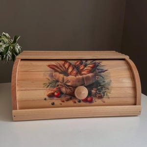 Può includere: Un portapane in legno con coperchio a rullo. La parte anteriore del portapane presenta un'immagine dipinta di un cesto pieno di pane, grano ed erbe. Il portapane è in legno chiaro e poggia su una superficie bianca.