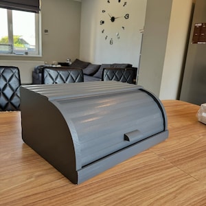 Puede incluir: Una panera de madera gris con tapa enrollable y un peque&ntilde;o asa. La caja rectangular tiene bordes redondeados y est&aacute; sobre una mesa de madera. La panera est&aacute; pintada en un color gris liso.
