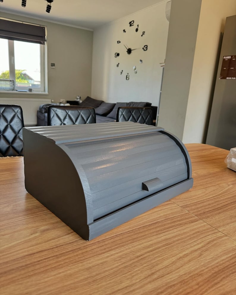 Puede incluir: Una panera de madera gris con tapa enrollable y un peque&ntilde;o asa. La caja rectangular tiene bordes redondeados y est&aacute; sobre una mesa de madera. La panera est&aacute; pintada en un color gris liso.