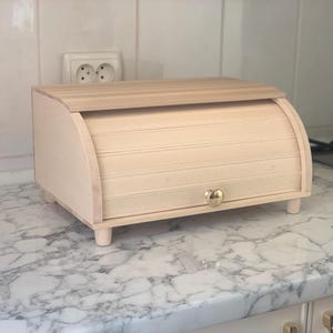 May include: A light-colored wooden bread box with a roll-top lid and a small, round, metallic knob. The rectangular container features a curved front panel and is supported by four short, cylindrical wooden legs, providing a stable base.