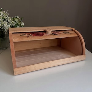 Op de afbeelding: Een lichtgekleurde houten brooddoos met een gebogen bovenkant en een scharnierend deksel. Het deksel heeft een geschilderd bloemenontwerp met rode rozen en groene bladeren. De doos is leeg en staat op een wit oppervlak.