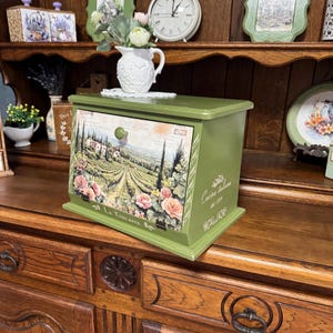 May include: A green wooden bread bin with a drawer featuring a Tuscan landscape scene with pink roses. The box has a round green knob and the words "La Toscana" and "Cucina Italiana" are visible.