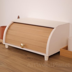 May include: A wooden bread box featuring a light beige roll-top door with a small, round, metallic gold knob. The frame of the box is painted white, and it stands on four short, sturdy white legs. The text ArtWoodPlanet is visible at the bottom.
