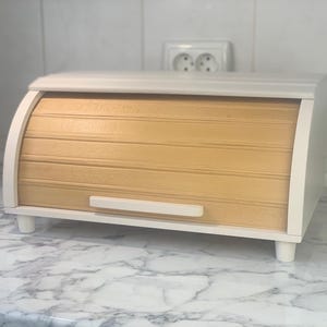 Op de afbeelding: Een houten broodtrommel met een roldeur. De trommel is gemaakt van lichtgekleurd hout met een natuurlijke afwerking en een wit frame. Het heeft een handvat aan de voorkant en staat op vier kleine, witte voetjes. De broodtrommel is ontworpen om brood en bakwaren in te bewaren.