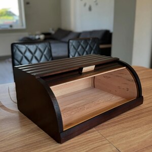 Puede incluir: Una panera de madera marr&oacute;n oscuro con un dise&ntilde;o curvo. La caja tiene una tapa de listones y un interior de color claro. La panera est&aacute; sobre una mesa de madera. El tema general es el almacenamiento de cocina.