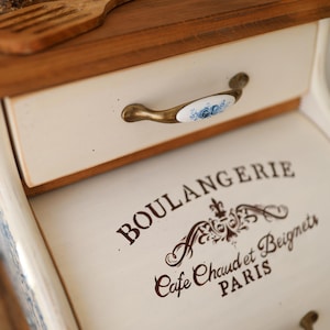 Puede incluir: Una panera de madera con un panel frontal de color crema que presenta el texto negro BOULANGERIE, Cafe Chaud et Beignets, PARIS. Tiene dos cajones con tiradores de tono lat&oacute;n acentuados por inserciones de cer&aacute;mica blanca con motivos florales azules.