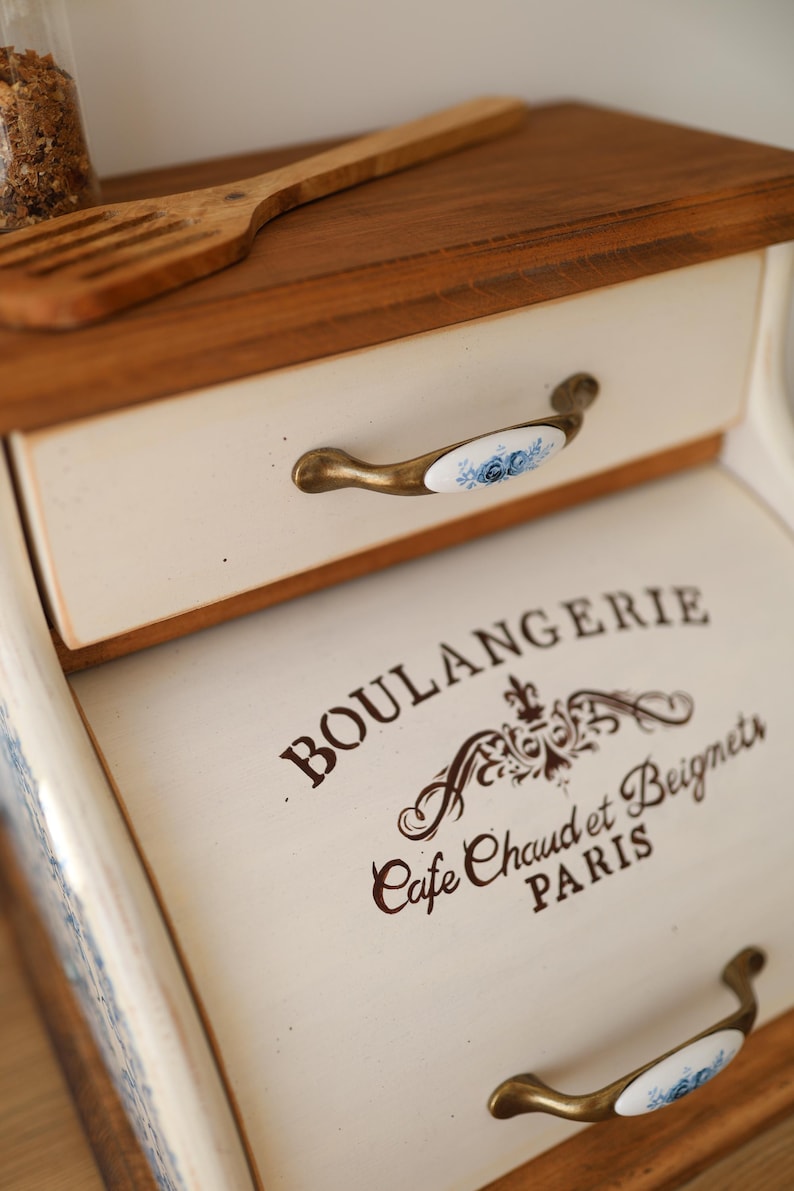 Puede incluir: Una panera de madera con un panel frontal de color crema que presenta el texto negro BOULANGERIE, Cafe Chaud et Beignets, PARIS. Tiene dos cajones con tiradores de tono lat&oacute;n acentuados por inserciones de cer&aacute;mica blanca con motivos florales azules.
