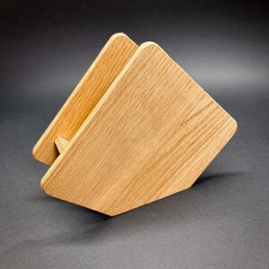 Puede incluir: Un servilletero de madera con acabado natural. El soporte est&aacute; hecho de dos piezas de madera en &aacute;ngulo unidas, creando una forma triangular. La veta de la madera es visible y el soporte est&aacute; sobre un fondo oscuro.