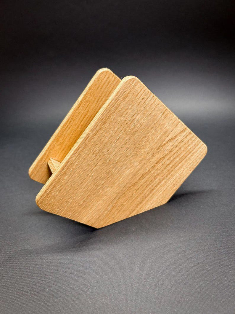 Puede incluir: Un servilletero de madera con acabado natural. El soporte est&aacute; hecho de dos piezas de madera en &aacute;ngulo unidas, creando una forma triangular. La veta de la madera es visible y el soporte est&aacute; sobre un fondo oscuro.