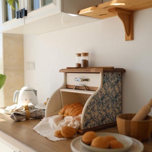 Puede incluir: Una panera de madera con puerta enrollable y paneles laterales con un patr&oacute;n floral azul y blanco. La unidad incluye un peque&ntilde;o caj&oacute;n superior con un tirador de metal y contiene pan rebanado y panecillos sobre un pa&ntilde;o blanco.