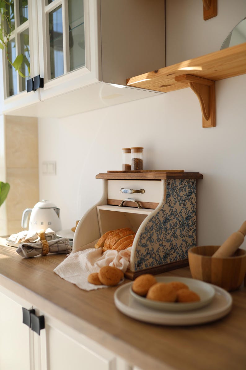 Puede incluir: Una panera de madera con puerta enrollable y paneles laterales con un patr&oacute;n floral azul y blanco. La unidad incluye un peque&ntilde;o caj&oacute;n superior con un tirador de metal y contiene pan rebanado y panecillos sobre un pa&ntilde;o blanco.