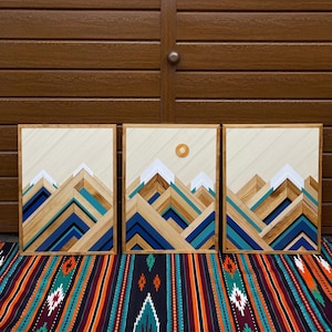 Op de afbeelding: Drie houten wandkunstwerken met een bergontwerp. De bergen zijn gemaakt van verschillende tinten blauw, wit en natuurlijke houttinten. Eén stuk heeft een houten zon. Het kunstwerk wordt getoond op een kleurrijk patroontextiel.