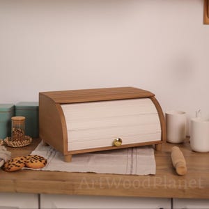 以下が含まれることがあります： ロールトップドア付きの木製ブレッドボックス。ボックスは薄茶色で、白のスラットフロントと金色のノブが付いています。2つの白いマグカップと麺棒など、他のキッチンアイテムと一緒に木製の表面に置かれています。