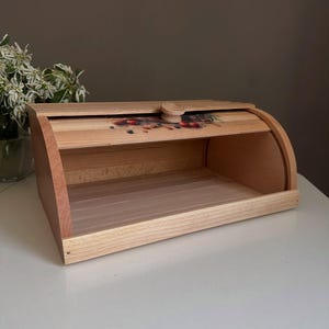 May include: Wooden bread box with a roll-top lid. The box is made of light-colored wood with a darker wood trim. The lid features a decorative floral design and a wooden knob. The bread box is empty and sits on a white surface.