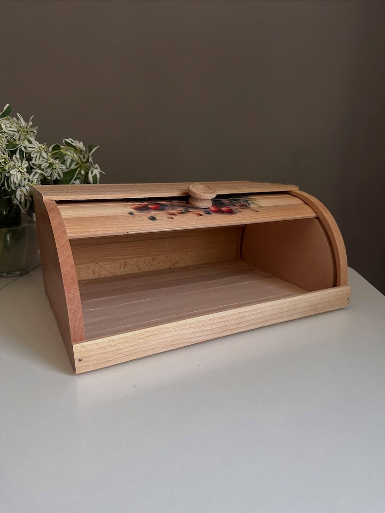 May include: Wooden bread box with a roll-top lid. The box is made of light-colored wood with a darker wood trim. The lid features a decorative floral design and a wooden knob. The bread box is empty and sits on a white surface.